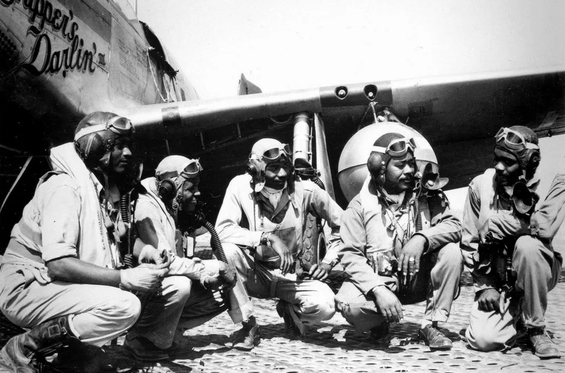 Archived black and white picture of five Tuskegee Airmen in conversation next to airplane